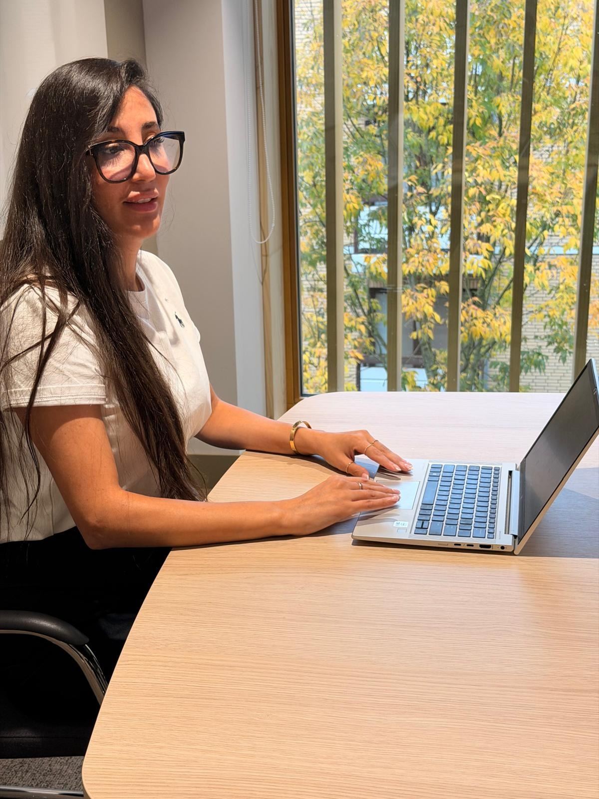 Rachida aan tafel met haar laptop, met herfstbomen achter het raam